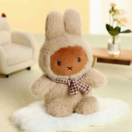 A plush toy rabbit wearing a checkered scarf sits on a white surface, with a white chair and a vase of flowers visible in the background.