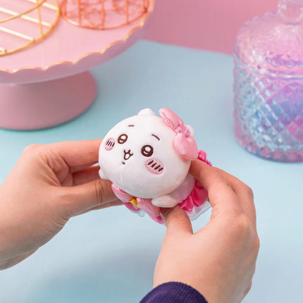 White plush mini mascot with a pink bow and ruffle trim, held in hands.