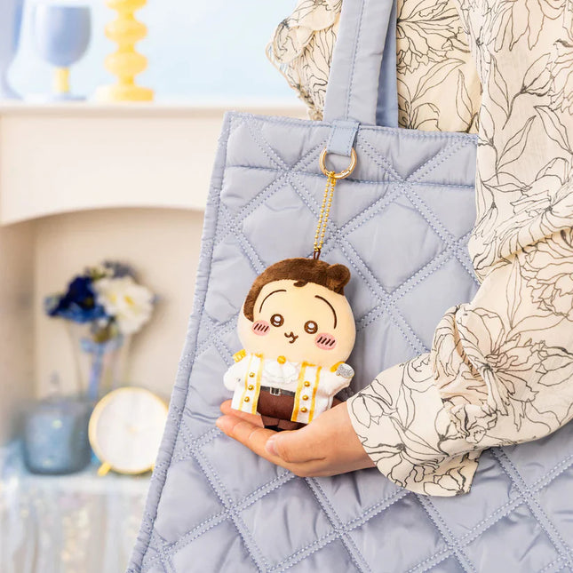 White plush mascot keychain with pink ruffled dress, held on gray tote.
